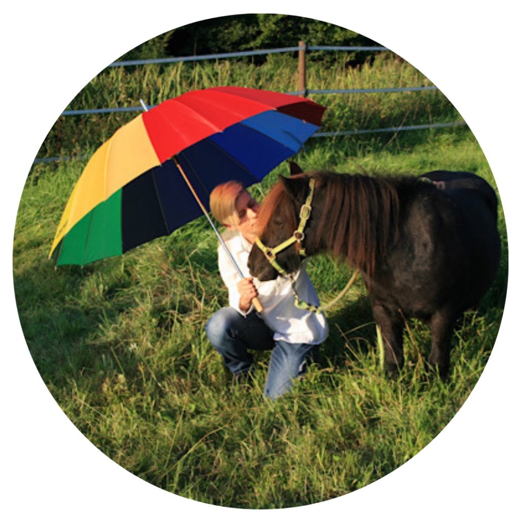 Frauke Porbeck, Mutter eines trans* Kindes kniet mit Regenbogen-Schrim neben einem Pony im Gras