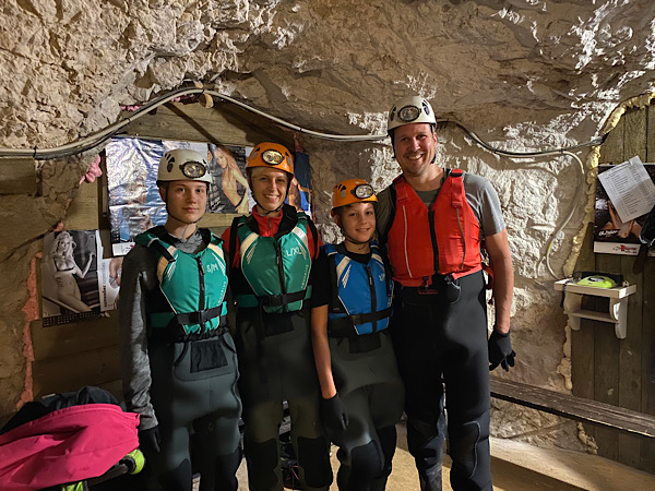 Vier lachende Menschen in Neoprenanzügen und Schwimmwesten mit Helmen und Kopflampen in einer Höhle